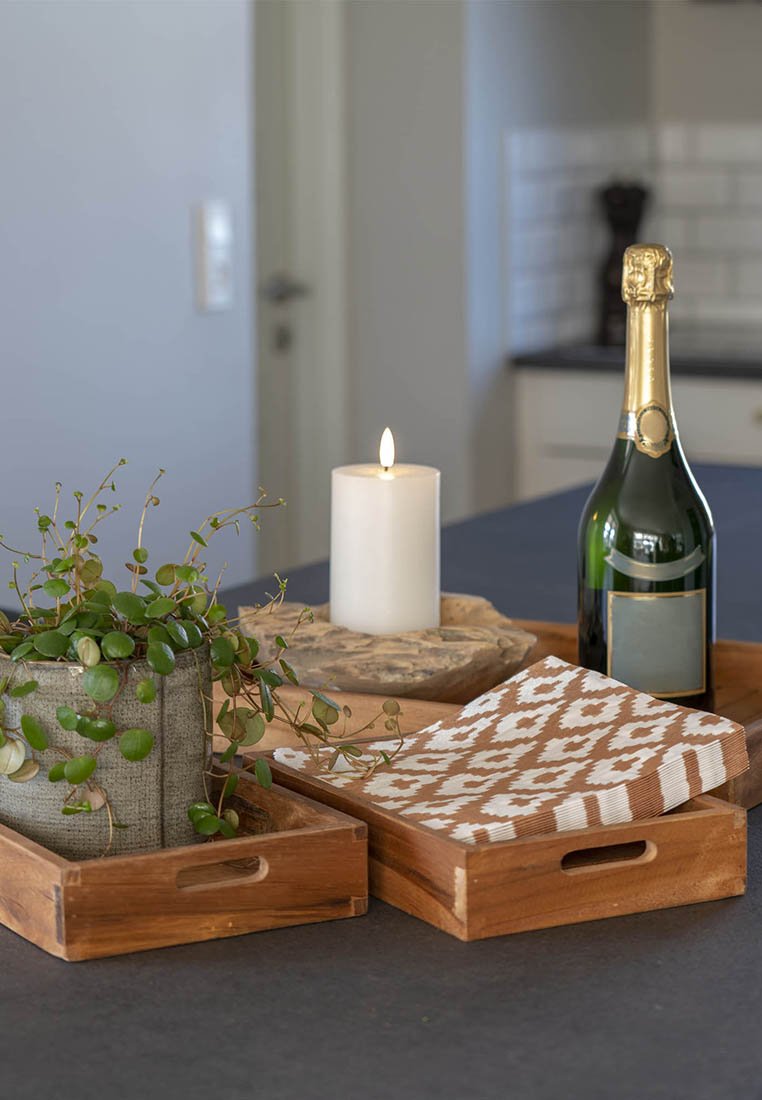 Holztabletts mit einem strukturierten Design, darauf eine weiße Kerze, Grünpflanzen in einem Stofftopf und eine Flasche Champagner auf einer dunklen Oberfläche.