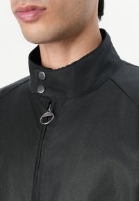 Black jacket with a stand collar, featuring snap buttons and a silver zipper pull. The fabric appears smooth and slightly glossy.