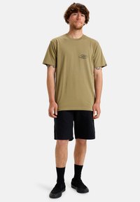 Camiseta de algodón verde oliva de manga corta, con un pequeño gráfico negro en el pecho; combinada con pantalones cortos negros y zapatillas negras sin cordones.