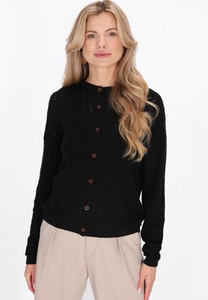 Lyshåret kvinde iført en sort knapret cardigan og beige bukser mod en ensfarvet hvid baggrund, vendt mod kameraet med et neutralt udtryk.