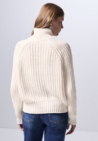 Chunky Creme Pullover mit hohem Kragen, gerippter Textur und überschnittenen Schultern. Trägt man zu dunkelblauen Jeans, die eine lockere Passform zeigen.