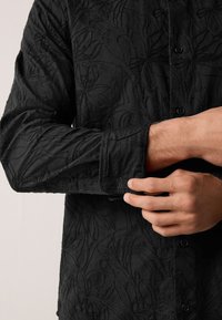 Schwarzes Hemd mit strukturiertem Blumenmuster, langen Ärmeln und Knopfleiste. Dezenter Glanz im Stoff sichtbar.