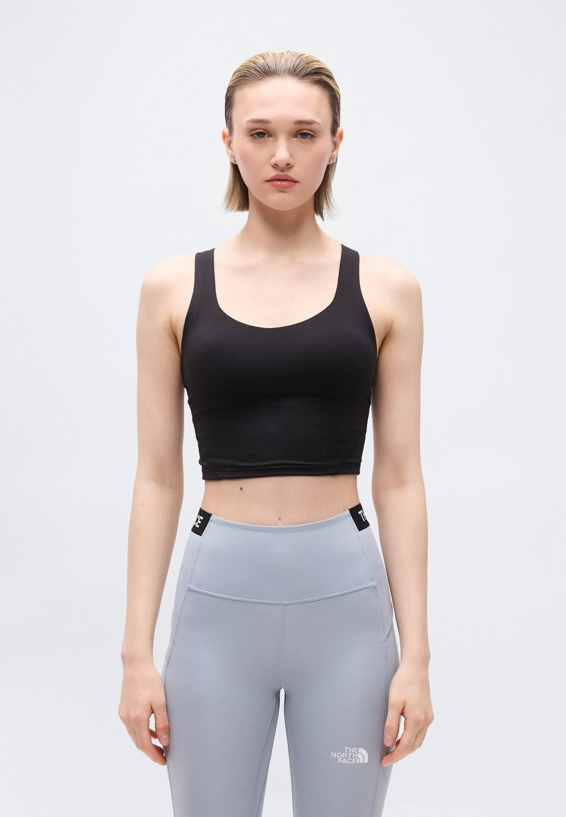 Noor naine lühikeste blondide juustega, kandes musta lühikest pluusi ja kõrge vöökohaga halli retuusid The North Face logoga, seistes ühevärvilise tausta ees.