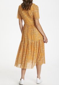 Mujer de pie con un vestido midi amarillo mostaza con pequeño estampado de flores blancas y zapatillas blancas, vista desde atrás.