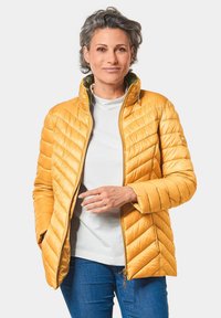 Veste matelassée jaune avec fermeture éclair, col montant et couture diagonale ; portée sur une chemise blanche et associée à un jean en denim bleu.