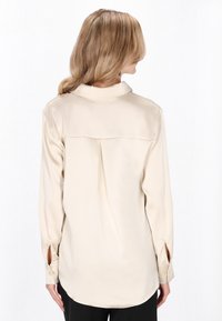 Vaaleanbeige silkkipaita, jossa on kaulus, pitkät hihat, sivuleikkaukset hihansuissa ja laskostettu takaosa.