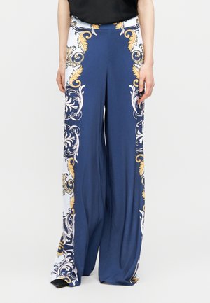 Pantalon large couleur bleu marine avec des motifs baroques ornés en or et blanc le long des côtés, porté avec un haut noir et des chaussures noires.