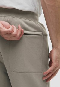 Close-up van een persoon die de ritssluiting van een zijzak van lichtgrijze joggingbroek met elastische tailleband omhoogtrekt.
