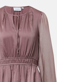Bluzka w kolorze mauve wykonana z lekkiego materiału, z dekoltem w serek, detalem w postaci wiązania, marszczoną talią oraz przezroczystymi długimi rękawami o subtelnej fakturze.