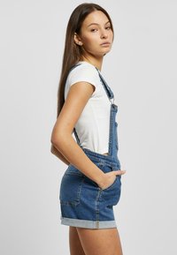 Denim overalls with a light blue wash, featuring adjustable straps, side pockets, and cuffed shorts. Worn over a white short-sleeve shirt.