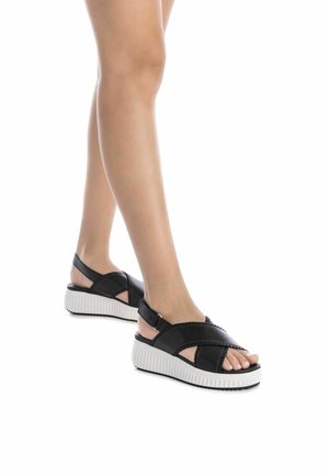 Sandalias de cuero negro con diseño de tiras cruzadas, correa ajustable en el tobillo y suela blanca estriada. Punta abierta con detalles de textura suave.