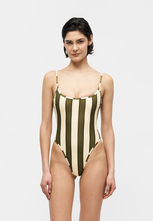 Femeie purtând un costum de baie întreg cu dungi verticale verde măsliniu și bej, bretele subțiri și croială înaltă pe picioare, stând în picioare în fața unui fundal simplu.