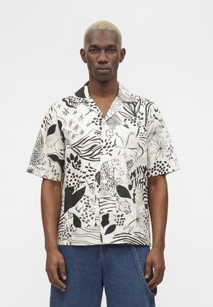 Mężczyzna o krótkich blond włosach, ubrany w czarno-białą, wzorzystą, krótkorękawową koszulę na guziki i niebieskie dżinsy na jednolitym tle.