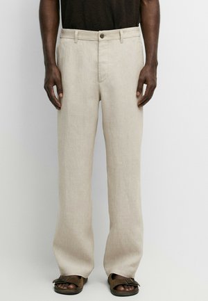Homme portant un pantalon en lin beige, une chemise marron foncé et des sandales brunes, debout devant un fond uni et clair, les bras le long du corps.