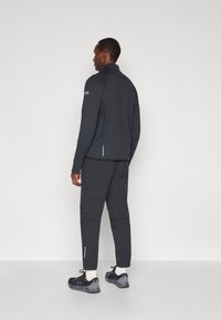 Melnā sportiskā jaka ar augstu apkakli un teksturētu audumu, apvienota ar atbilstošām kravas biksēm un melniem apaviem ar pelēkiem akcentiem.