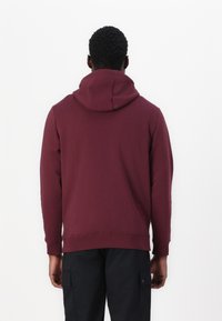 Sweat à capuche bordeaux avec une grande capuche, des manches longues et des poignets côtelés. Fabriqué en tissu doux et texturé, avec une silhouette ajustée, associé à un pantalon noir.