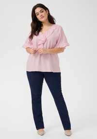 Blusa rosa chiaro con collo arricciato e maniche corte, abbinata a jeans in denim blu scuro. Il tessuto sembra leggero e leggermente strutturato.