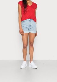 T-shirt rossa con scollo a V e maniche corte, abbinata a pantaloni corti di jeans blu chiaro con risvolti, indossata con sneakers bianche con piattaforma.