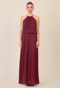 Burgundy pleated maksi dresses dengan desain leher halter, menampilkan kain tekstur dan siluet mengalir. Hem sederhana dan sepatu datar terlihat.
