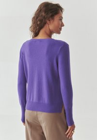 Maglione viola con maniche lunghe, scollatura a costine e orlo a costine. Tessuto morbido, design aderente e spacchi laterali. Abbinato a pantaloni beige.