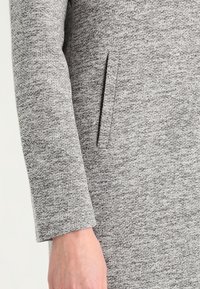Grau gestrickter Jackenstoff mit feiner Textur, ausgestattet mit einer Seitentasche und einem geraden Ärmelabschluss.