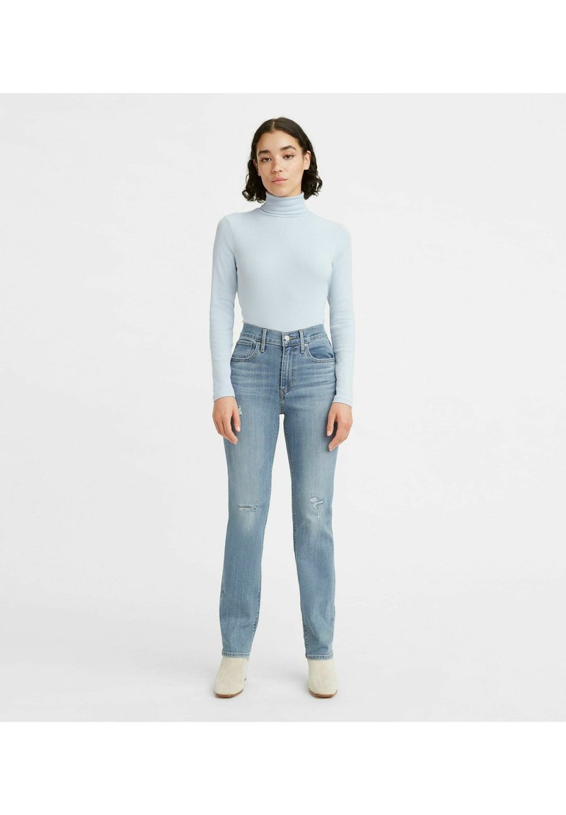 Mujer de pie contra un fondo blanco, vestida con un suéter de cuello alto azul claro, unos jeans ajustados de talle alto con leve desgaste, y botas blancas hasta el tobillo.