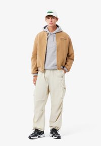 Giacca bomber di lana marrone sopra un hoodie grigio, pantaloni cargo color crema e sneakers bianco/nero. Cappello bianco con logo di un coccodrillo verde.