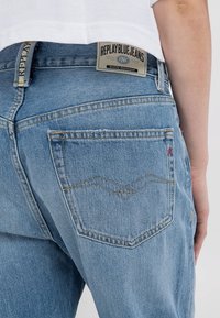 Bakifrån av en person som bär ljusblå Replay jeans och en vit skjorta, som visar ficksömmar och varumärkesetikett vid midjan.