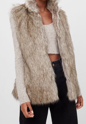 Femme portant un gilet en fausse fourrure beige sur un crop top à manches longues beige clair et un pantalon noir taille haute.