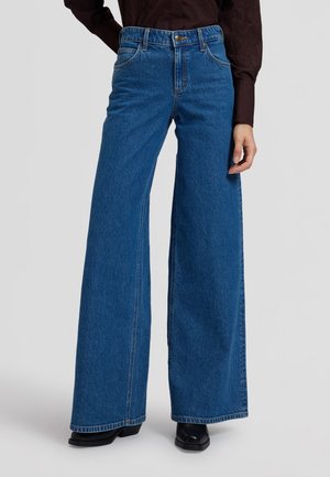 Personne portant un jean bleu taille haute à jambes larges avec poches avant, des chaussures noires à talons et une chemise sombre à manches longues sur un fond uni.