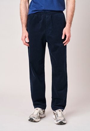 Man draagt marineblauwe ribfluwelen broek, witte en grijze hardloopschoenen en een blauw overhemd tegen een effen lichte achtergrond.