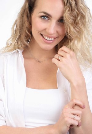 Collier en argent avec un pendentif en forme de cœur porté sur le cou d'un mannequin, associé à une fine bague en argent ; le mannequin a des cheveux blonds ondulés et porte une chemise blanche.