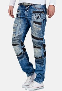 Jeans in denim blu chiaro con accenti in tessuto nero, multiple tasche, dettagli di design cuciti e motivi testurizzati.