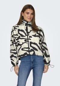 Giacca cropped in un materiale beige lucido con motivi astratti neri, dotata di un alto colletto e polsini elasticizzati. Chiusura con zip sul davanti.