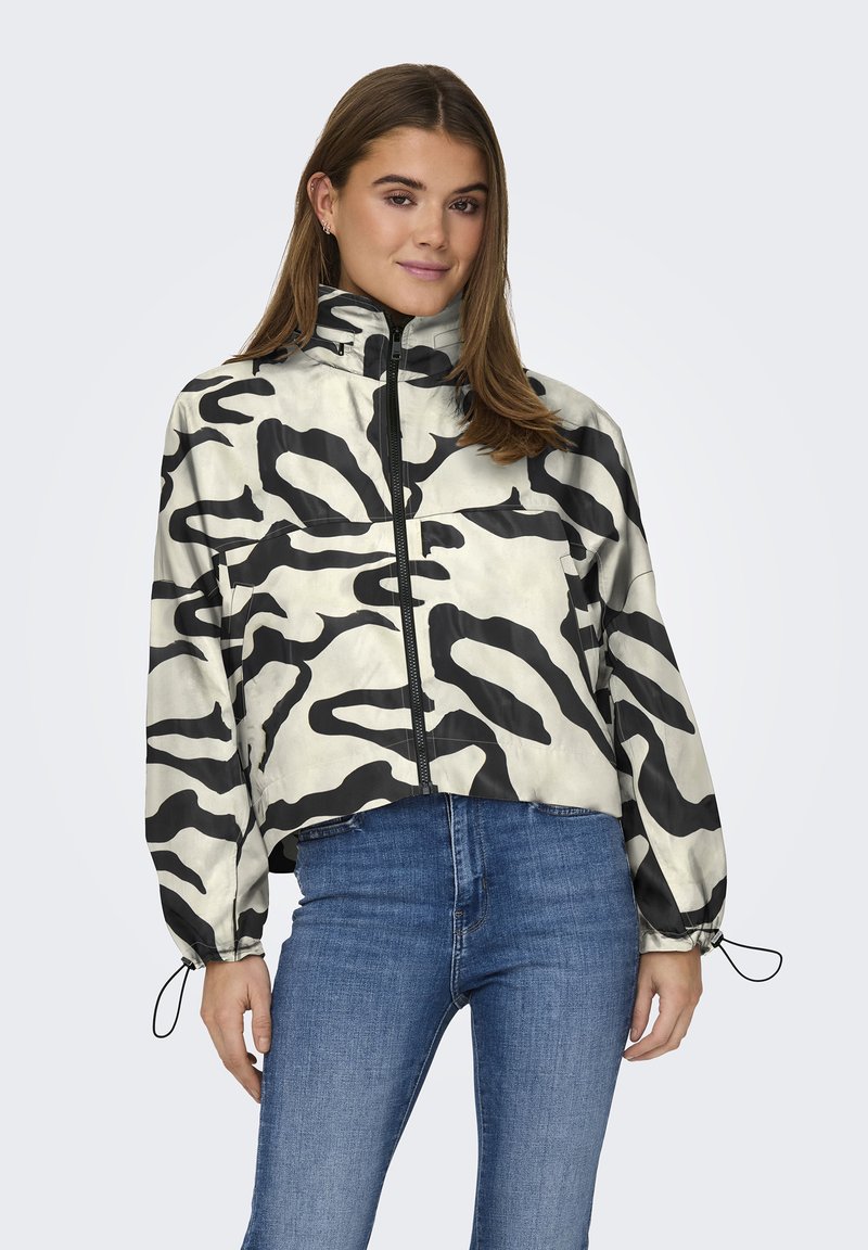 Giacca cropped in un materiale beige lucido con motivi astratti neri, dotata di un alto colletto e polsini elasticizzati. Chiusura con zip sul davanti.