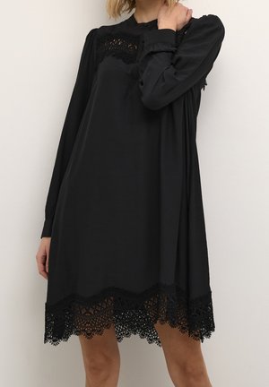 Frau trägt ein langärmeliges schwarzes Kleid mit Spitzendetails an Brust und Saum, steht vor einem schlichten weißen Hintergrund.