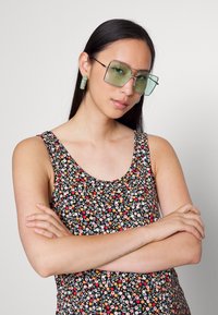 Mulher com cabelo longo e escuro, usando óculos de sol quadrados oversized com tonalidade verde, uma blusa floral sem mangas e brincos retangulares verdes, com os braços cruzados.