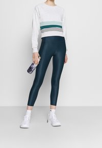 Zkrácená světle šedá mikina s pruhy v barvách tyrkysová, tmavě modrá a šedá, doplněná tmavě tyrkysovými legínami a bílými teniskami, držící lahvičku s vzorem.