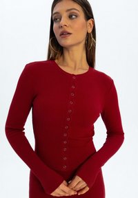 Czerwona, prążkowana bluzka z długim rękawem, prezentująca okrągły dekolt i zapięcie na guziki z metalowymi akcentami z przodu. Dopasowany krój z przedłużonymi mankietami.