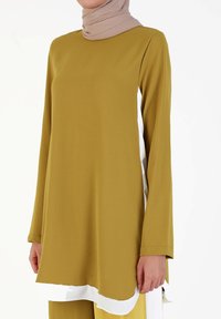 Olive green long-sleeve tunic with a curved hemline and contrasting white side panels, made of soft fabric. Paired with beige headscarf.