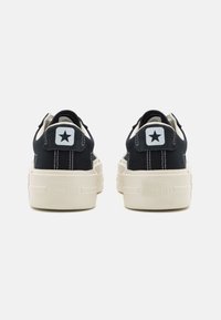 Black canvas sneakers with a white rubber sole, featuring a star logo on the heel and contrasting stitching. Low-profile design with tab detail.