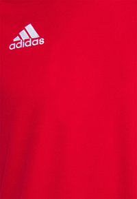 Piros sportos póló sima anyagból, a bal felső oldalon fehér Adidas logóval. Egyszerű, rövid ujjú tervezés.