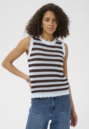 Femme aux cheveux bouclés vêtue d'un haut tricoté sans manches rayé bleu et marron et d'un jean bleu taille haute, debout avec une main sur la hanche.