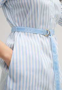 Camicia a righe blu chiaro e bianca, dotata di una cintura abbinata con un anello dorato e dettagli del logo. Include tasche laterali e chiusura a bottoni.