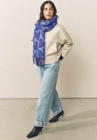 Sciarpa blu con motivo a cuori, giacca beige, jeans azzurri e stivaletti neri con tacco. La sciarpa presenta frange alle estremità.