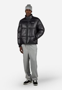 Svart puffjakke med høy krage og blank tekstur, kombinert med lysegrå bukser og svarte joggesko. Strikket grå lue på hodet.