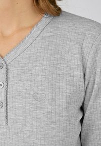 Top gris de manga larga con canalé y escote en V, con botones decorativos y un pequeño diseño de corazón bordado en el pecho.
