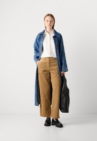 Vrouw staat gekleed in een lange spijkerjas, wit overhemd met knopen, tan kleurige corduroy broek, zwarte loafers en houdt een grote zwarte tas vast.