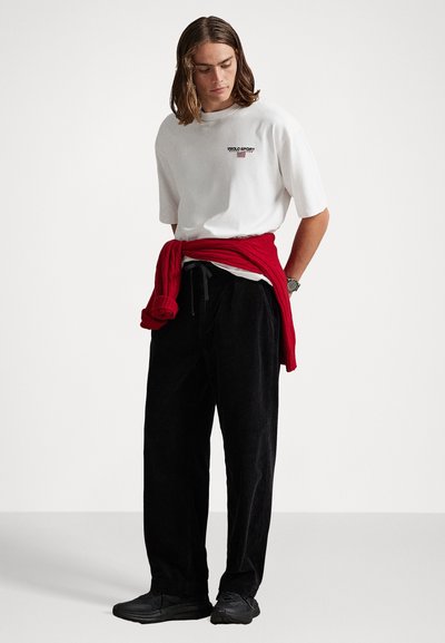 Hombre joven con cabello hasta los hombros, vistiendo una camiseta blanca de Polo Sport, pantalones negros, zapatos negros y un suéter rojo atado a la cintura.
