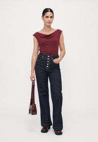 Mujer vestida con una blusa burdeos y jeans oscuros de cintura alta, sosteniendo un bolso burdeos a juego, de pie con una mano en el bolsillo.